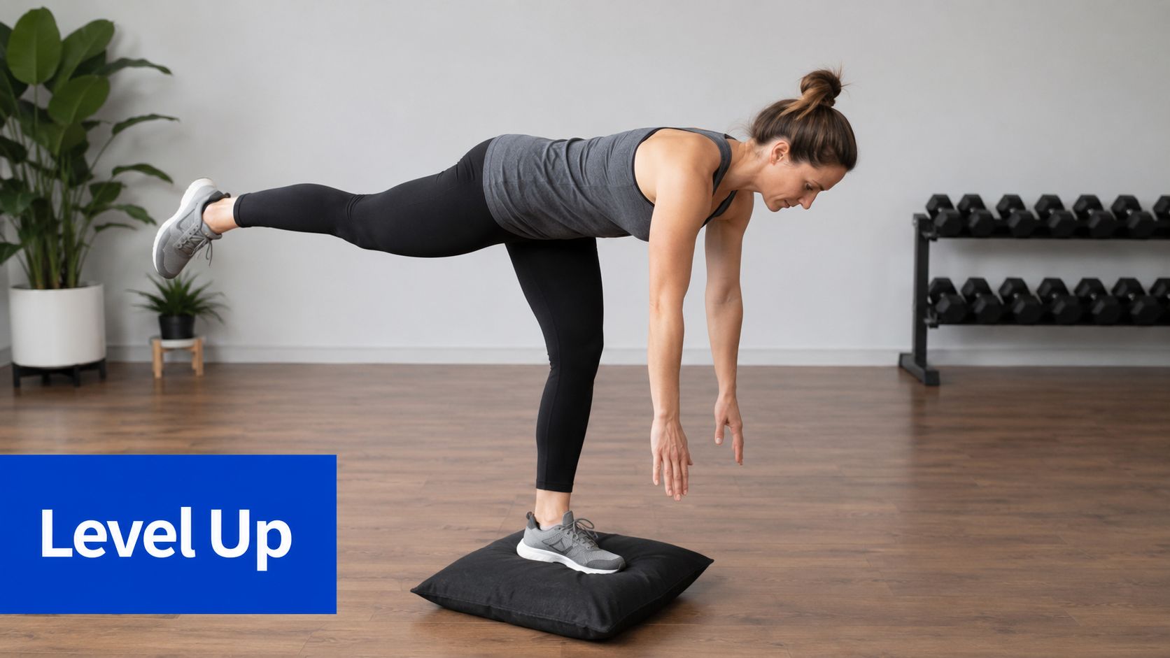 A woman performing a single leg balance exercise on a black cushion in a gym studio.
