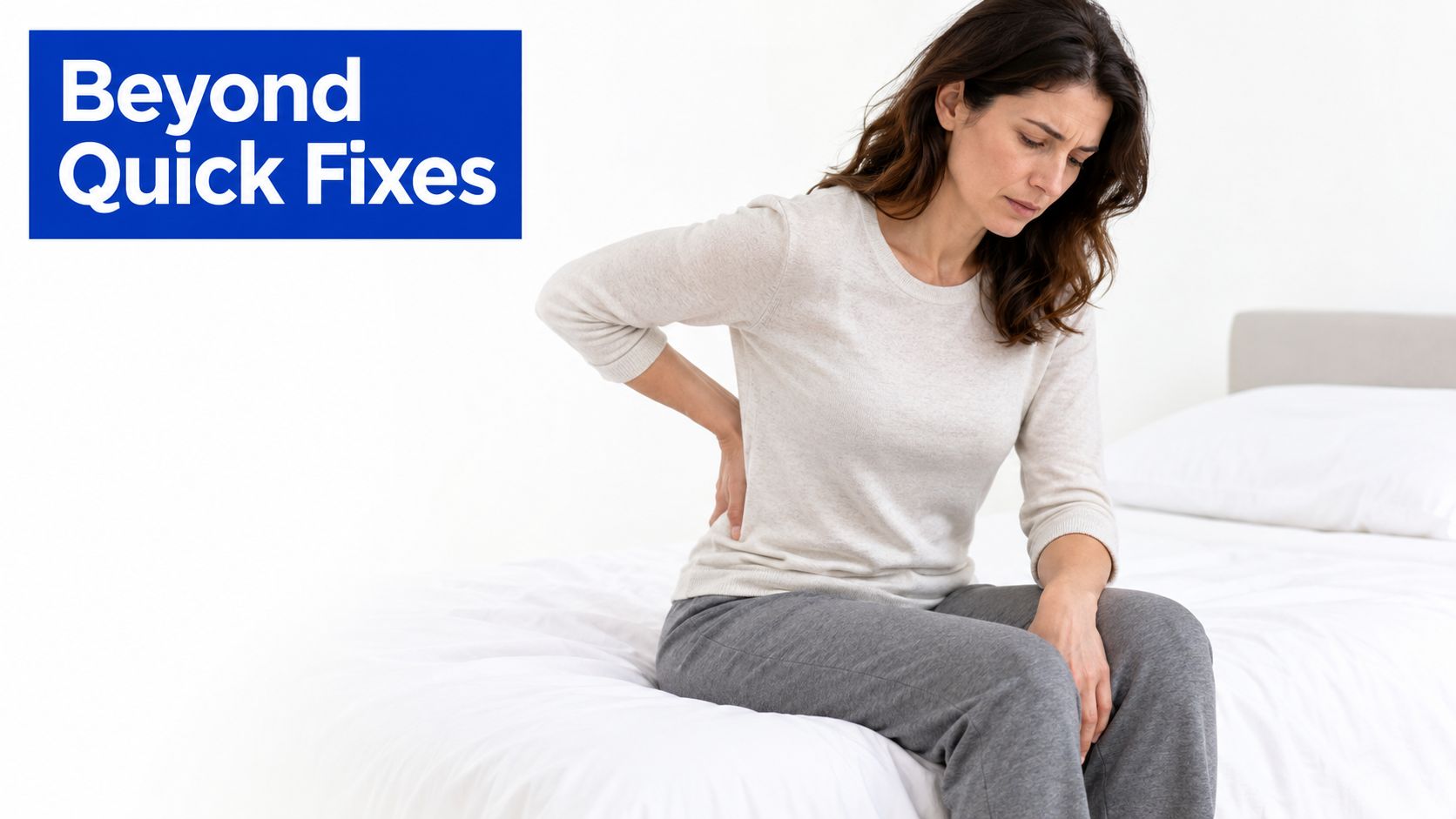A woman sitting on a bed looking distressed while holding her lower back in pain and discomfort.